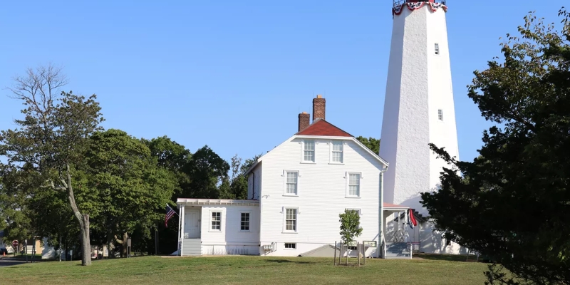 Sandy Hook Lighthouse, a white structure with a red roof, is surrounded by a grassy field, highlighting nearby beaches.