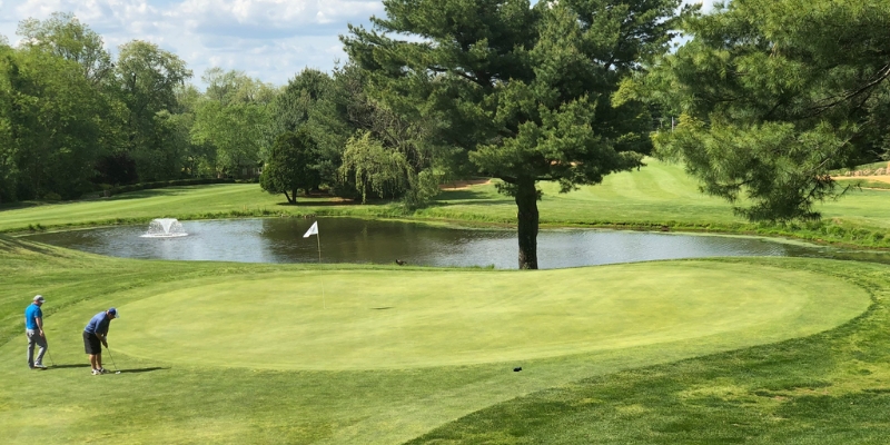 Two men playing golf on a green course at Bella Vista Country Club, with water visible in the background.