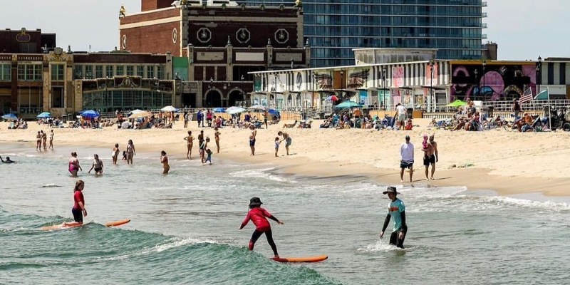 People surf at Asbury Park Beach, with the boardwalk and a nearby building creating a lively beach atmosphere.
