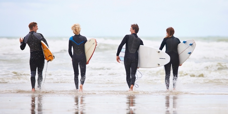Four surfers in wetsuits stroll on Bradley Beach, each holding a surfboard as they head towards the water.