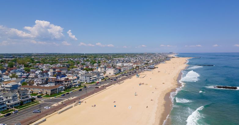 Aerial view of a wide New Jersey beach with homes, a road, and gentle waves under a clear blue sky with scattered clouds.


