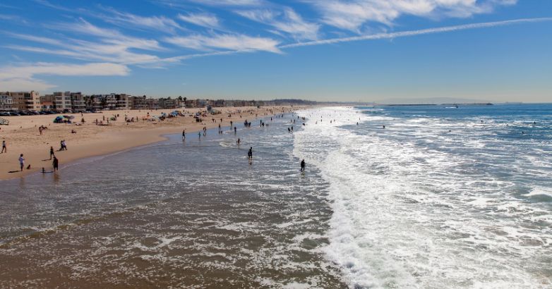 Busy New Jersey beach with people in the surf, sandy shore, beachfront homes, and a partly cloudy sky above the ocean.






