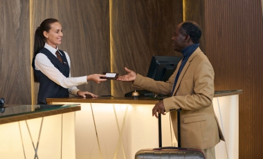At the hotel front desk, a man gives a woman a piece of paper, highlighting concierge services for Seastreak to NYC.