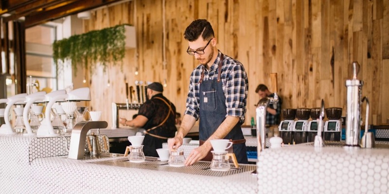 A man in an apron brews coffee at a counter, showcasing local coffee shops.