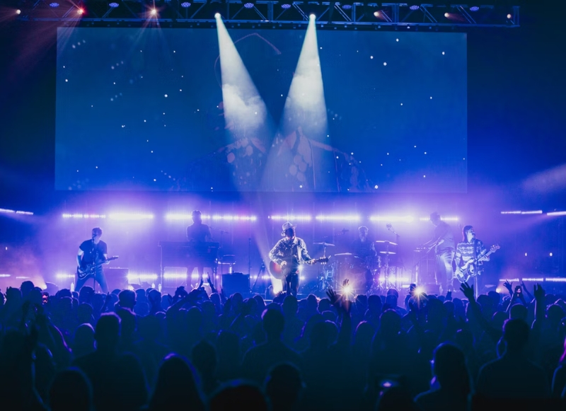 A lively concert scene featuring performers on stage with a large screen displaying visuals behind them.