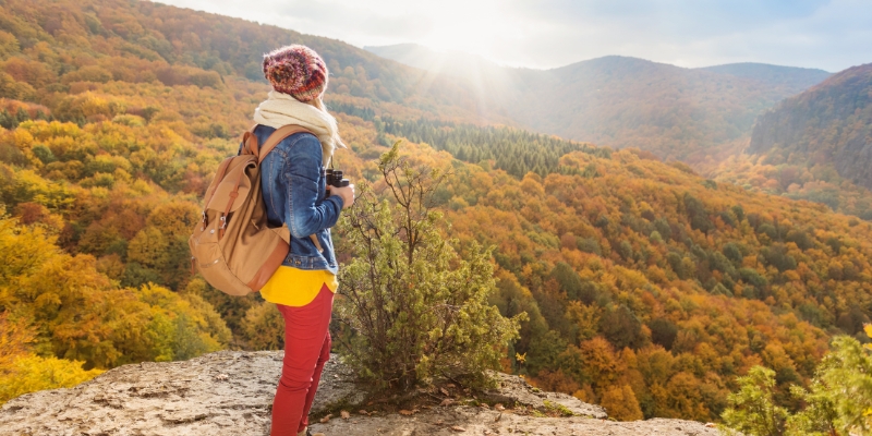 Woman with a backpack stands atop a mountain, gazing at the vibrant autumn forest in New Jersey.