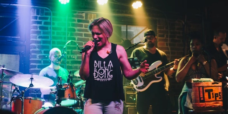 A woman performs on stage, singing passionately with a band accompanying her in a vibrant music setting.