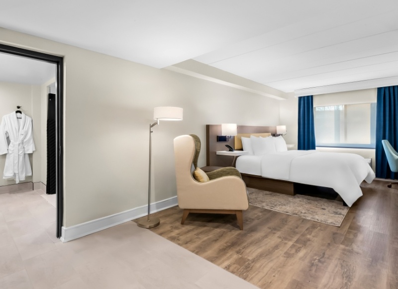 Modern hotel suite with a king bed, wingback chair, and view into the bathroom.