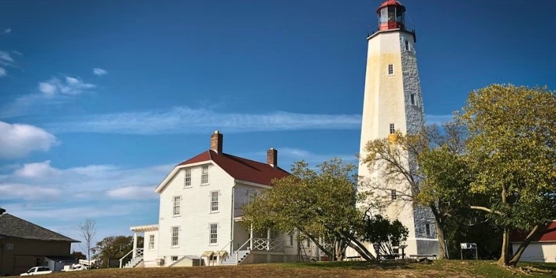 A lighthouse on a hill beside a white house, set against the backdrop of Sandy Hook at Jersey Shore.