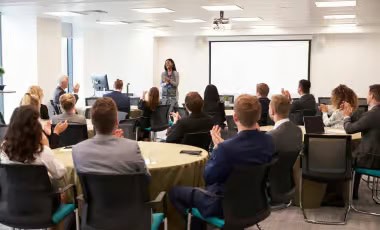 Audiovisual Support Audience sitting at round tables applauding a speaker presenting in a bright conference room.