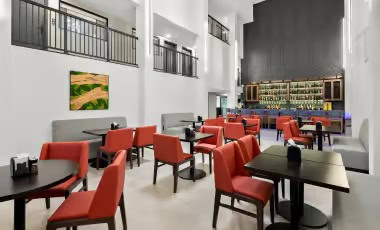 On-Site Dining Spacious hotel lounge and dining area with red chairs, dark tables, and a bar in the background.