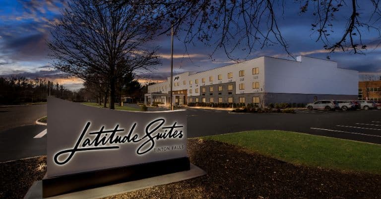 Exterior view of Latitude Suites Tinton Falls hotel with an illuminated sign at dusk.